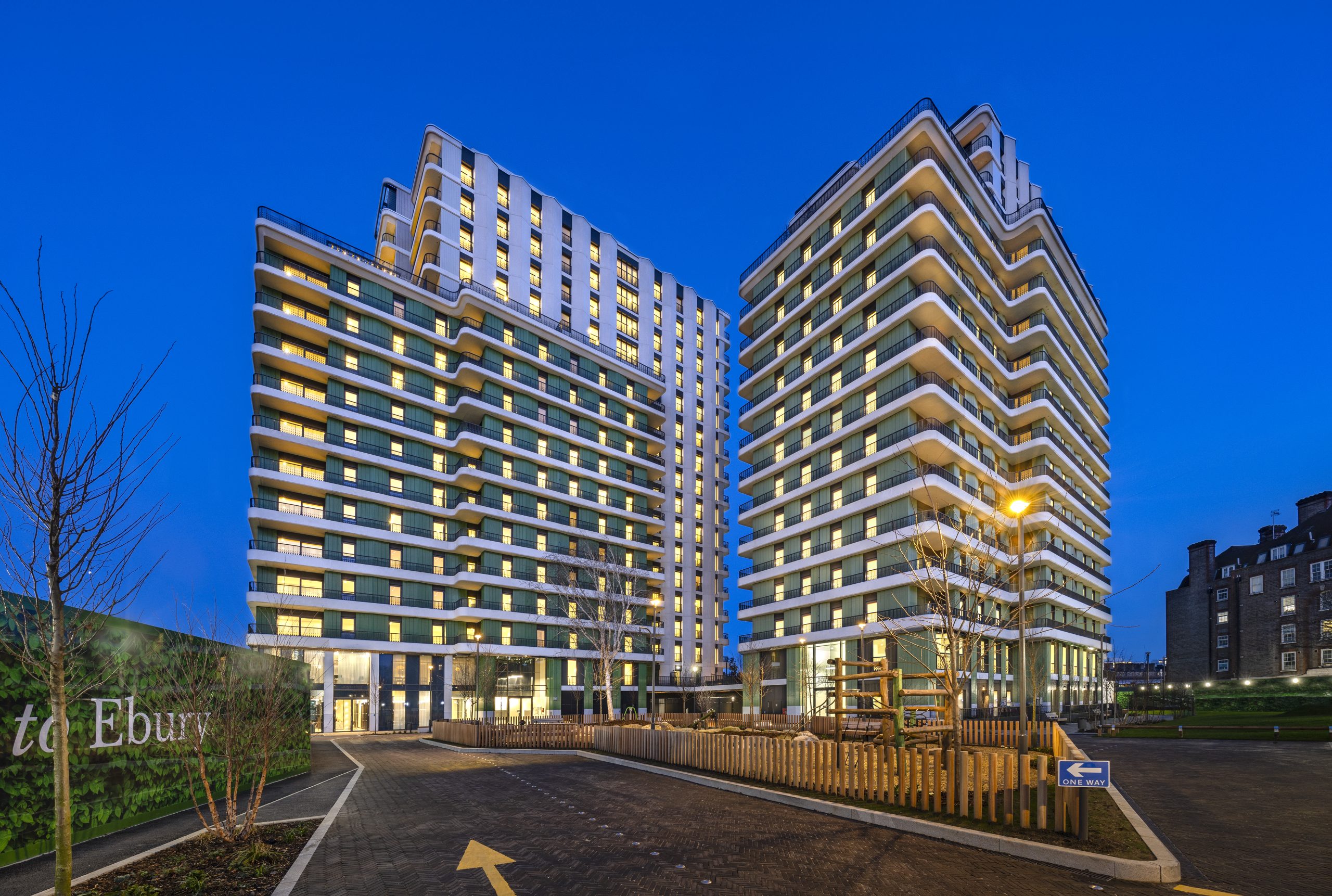 The Ebury development as viewed from the outside street in the evening. We see two buildings with the lights on in all windows as well as the road leading towards them